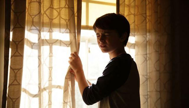 Preteen squinting while pulling curtains backlit by morning light through patterned drapes