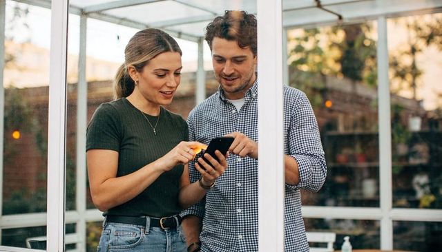 Young couple sharing smartphone in glass conservatory, browsing together in sunlit space