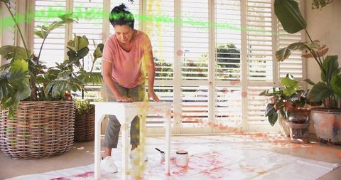 Mature woman sanding white wooden table in sunroom amid plants diy furniture restoration
