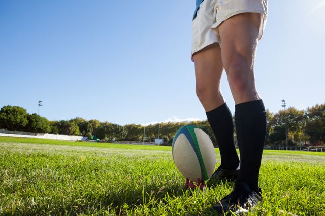 Low section of player standing by rugby ball on playing field against clear blue sky