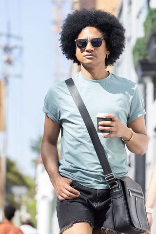 African american man strolling city sidewalk holding coffee wearing crossbody bag and sunglasses