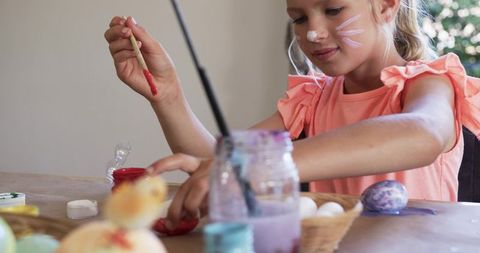 Young girl painting Easter eggs at table with face paint and colorful paint jars
