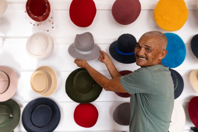 Portrait of a happy senior biracial man smiling and showing a hat surrounded by multiple colorful hats on display in the showroom at a hat factory.