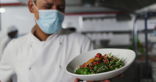 Chef showing freshly prepared vegetable salad wearing face mask in a professional kitchen. Ideal for highlighting industrial culinary environments, food hygiene standards, and healthy dining options in restaurants. Perfect for use in publications about kitchen health safety, chef lifestyles, or healthy eating.