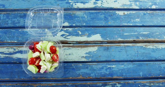 A fresh salad with cherry tomatoes and cucumbers is packed in a clear plastic container on a rustic blue wooden table, with copy space. The image evokes a concept of healthy eating on-the-go or meal prepping.