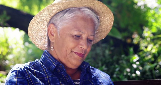 Senior woman with white hair wearing casual blue plaid shirt and straw hat is reading in a leafy garden. Ideal for campaigns promoting senior well-being, outdoor activities, and nature benefits for the elderly.