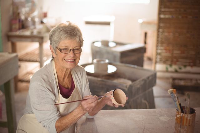 Senior woman painting a ceramic bowl in a pottery workshop. She is smiling and wearing glasses and an apron. The background shows various pottery tools and equipment. Ideal for use in articles about hobbies, senior activities, creative arts, and artisan crafts.