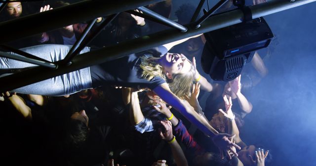 Young woman crowd surfing during a concert in a club environment. Audience members hands raised in support and excitement. Blue stage light creating dynamic atmosphere. Perfect for advertisements promoting music festivals, live events, parties, nightlife, and youth culture.