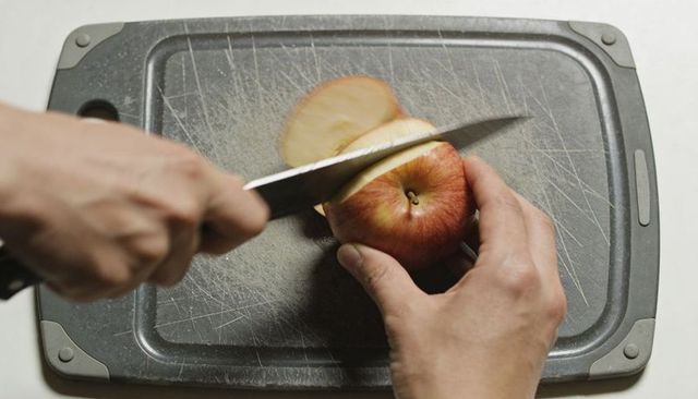Hands slicing red apple on scratched cutting board with knife overhead closeup food prep