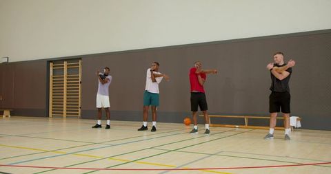Diverse male teammates stretching arms on indoor basketball court preparing for practice