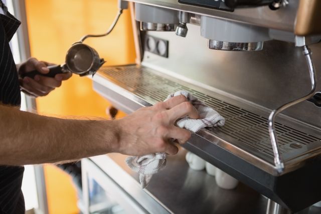 Waiter cleaning coffee machine in café, focusing on hygiene and maintenance. Ideal for use in articles or advertisements related to coffee shops, barista training, hospitality industry, and workplace cleanliness.