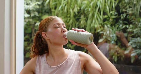 Fit woman drinking from textured bottle after home workout near sunlit window garden