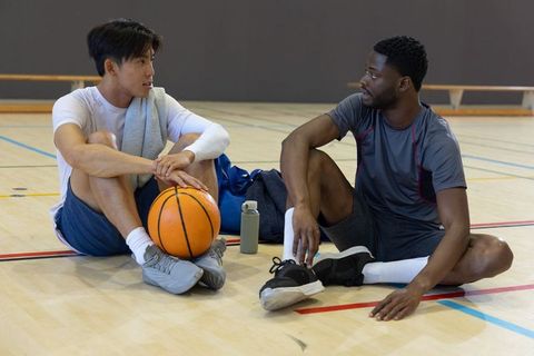 Asian and african american teammates sitting and talking with basketball on gym court