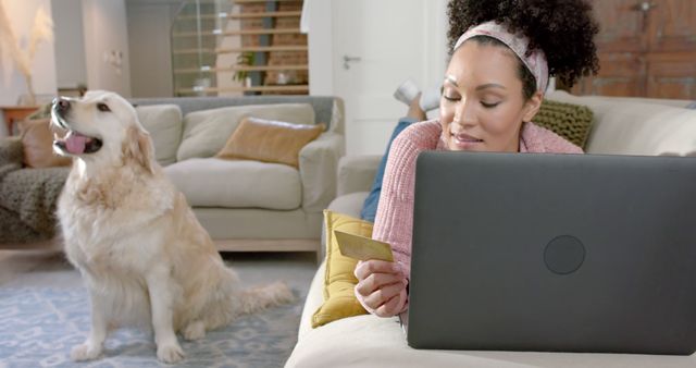 Woman using laptop and credit card while shopping online at home. A golden retriever dog is sitting near her, providing a comfortable and cozy atmosphere. Ideal for depicting home shopping experiences, leisure, comfort, pet companionship, and modern lifestyle. Suitable for use in advertisements, websites, blogs, and articles about e-commerce, home activities, and pet-friendly environments.