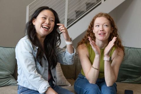 Two friends laughing on couch wearing smartwatches sharing cozy conversation at home