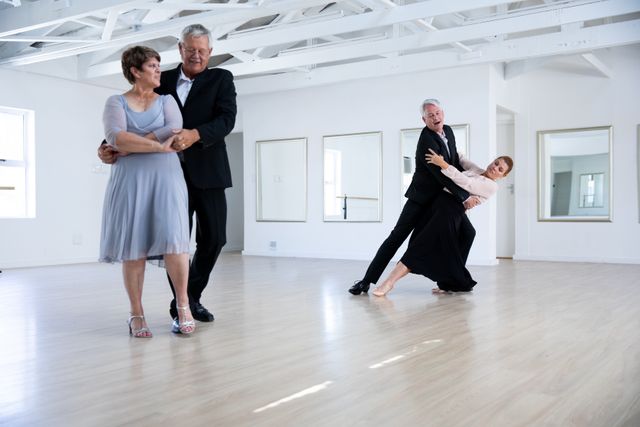 Two happy elegant Caucasian senior couples spending time together in a ballroom, taking part in dancing class, dancing, holding hands and smiling. Active golden years hobby.