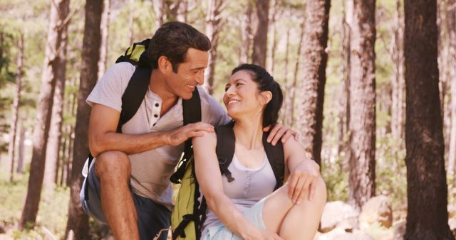 This image depicts a happy couple sitting in a forest, taking a break during a hike. They are both wearing backpacks and looking at each other with smiles. Perfect for use in travel brochures, adventure blogs, outdoor activity promotions, and relationship articles focused on spending quality time together.