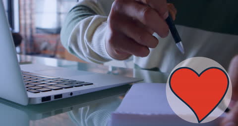 Close-Up Hand Writing with Laptop and Heart Icon Overlay