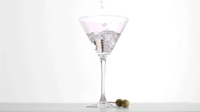 A martini is being poured in super slow motion into a classic clear glass with two green olives nearby against a minimalist white background. Perfect for use in advertisements for bars, restaurants, or liquor brands. It can also be used in articles, tutorials, or blogs related to cocktail making, mixology, and entertaining.