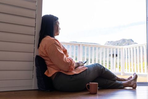 Mature woman sitting on sunlit balcony floor holding smartphone and coffee mug, serene