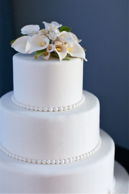 This elegant three-tier white wedding cake features a delicate floral topper and simple bead decoration. Ideal for use in wedding-related content, celebrations, party invitations, bakery advertisements, and festive occasions.