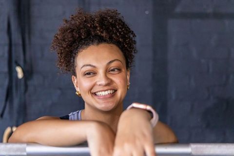 African american woman smiling and leaning on gym bar, curly bun and smartwatch, urban brick backgro