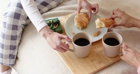Female friends sharing coffee and croissants on bed in plaid pajamas, cozy intimate morning