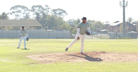 Right-handed pitcher delivering pitch with outfielder waiting on sunny community field
