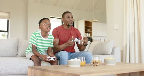 African american father and son playing video games on sofa with controllers and snacks