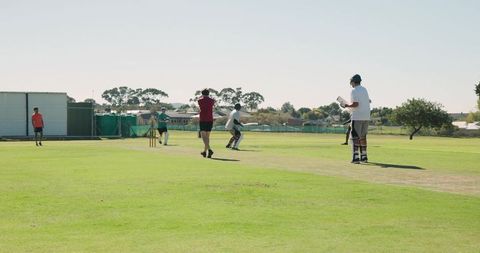 Suburban cricket match bowling delivery over trimmed pitch with batsman and fielders