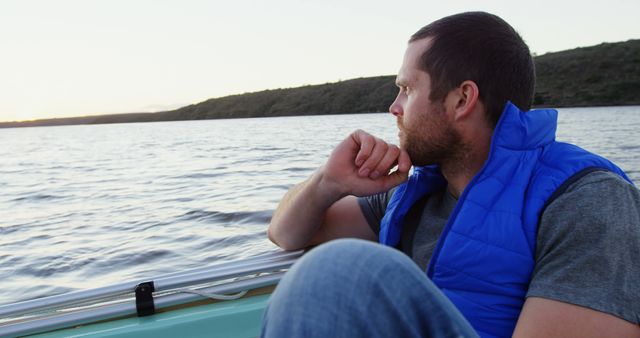 Man sitting on boat, looking at serene lake during sunset, wearing blue vest. Perfect for illustrating themes of relaxation, contemplation, and nature. Can be used for outdoor lifestyle, travel, and wellness content.