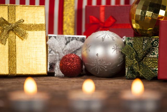 Christmas gift boxes and baubles arranged on a wooden table with lit candles in the foreground. Ideal for holiday greeting cards, festive advertisements, seasonal blog posts, and social media content celebrating Christmas and the holiday season.
