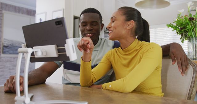 Image of happy diverse couple watching tablet together and laughing sitting in kitchen. Communication, happiness, love, domestic life and inclusivity concept.