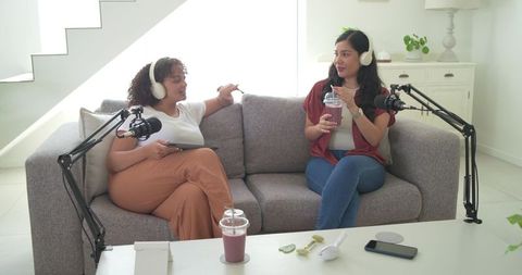 Diverse female cohosts recording casual podcast on sofa with smoothies and microphones