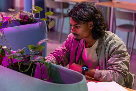 Indian man tending planter vines in modern coworking lounge with notebook and smartwatch