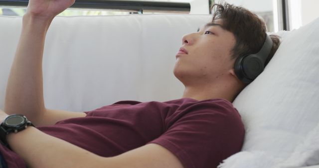 Asian male teenager wearing headphones and using smartphone in living room. spending time alone at home.