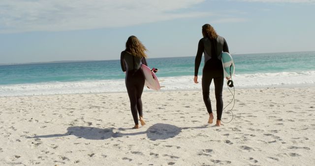 Two people wearing wetsuits walking on sandy beach towards ocean waves holding surfboards. Perfect for use in travel promotions, surfing equipment advertisements, beach tourism campaigns, and outdoor sport-themed blogs.