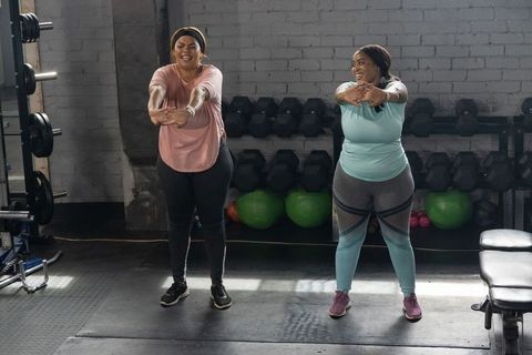 Two diverse women stretching and laughing during gym workout by dumbbell rack and medicine balls