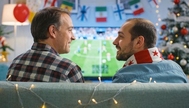 Middle-aged friends watching soccer on couch with festive holiday lights and scarf