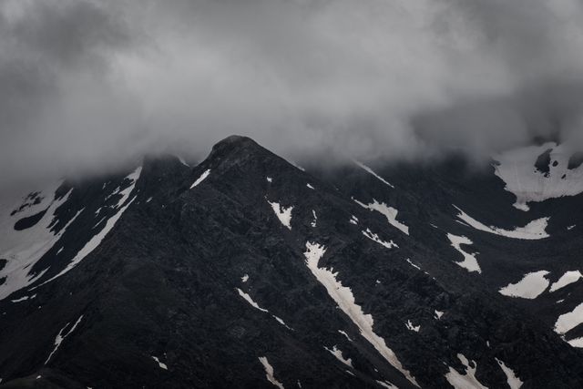 This image shows an atmospheric scene featuring dark, rugged mountain peaks partially covered in snow patches with a cloudy sky and mist hovering over the summits. It evokes a sense of mystery and wilderness, making it suitable for use in travel advertisements, adventure blogs, background images for websites, and material emphasizing natural beauty and remote, wild landscapes.