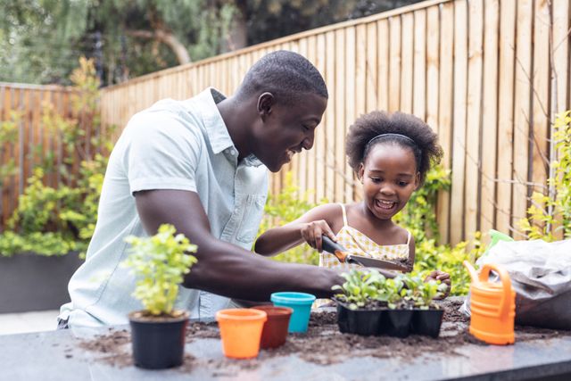 Happy african american father with excited daughter planting small potted plants in yard. Unaltered, family, togetherness, childhood, gardening and nature concept.