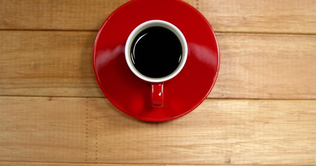 A red coffee cup filled with black coffee sits on a saucer atop a wooden table, with copy space. The vibrant contrast of the red against the wood grain provides a warm, inviting atmosphere for a coffee break.