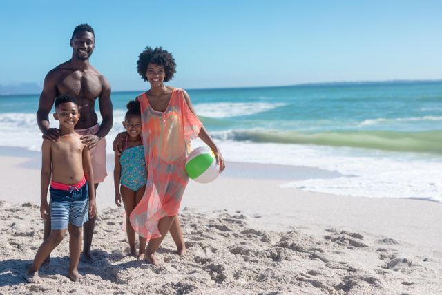 This image shows a joyful African American family of four enjoying a sunny day at the beach. The parents and their two children are smiling and standing on the sandy shore with the ocean in the background. The mother is holding a beach ball, indicating they are ready for some fun activities. This image can be used for promoting family vacations, summer activities, travel destinations, and lifestyle blogs.