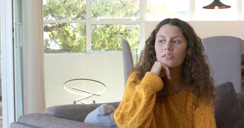 Pensive young woman sitting on gray sofa wearing mustard sweater in bright modern living room