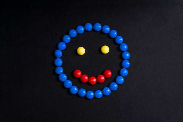 This image shows a smiling face creatively arranged using colorful candies on a black background. The candies form the outline of the face in blue, eyes in yellow, and smile in red. It is perfect for themes related to creativity, fun, joyful designs, children’s parties, confectioneries, greeting cards, and advertisements focusing on playful and cheerful concepts.