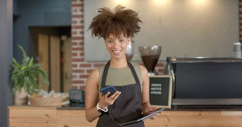 Smiling barista holding phone and tablet while working behind espresso counter