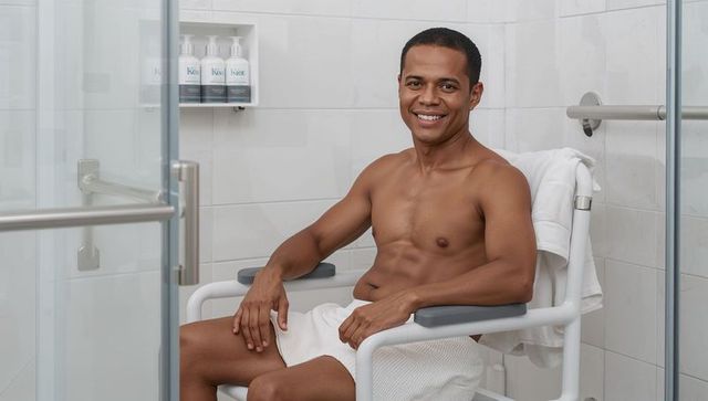 Smiling man sitting on shower chair in accessible bathroom with grab bars and glass door