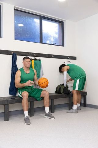 Basketball teammates resting and preparing on bench in bright locker room with ball