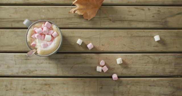 Top view of a mug filled with hot cocoa and pink and white marshmallows on wooden table. Brown autumn leaf beside the mug and scattered marshmallows add to the fall theme. Perfect for marketing campaigns, social media posts, websites, and blog articles about autumn comforts, seasonal drinks, or rustic lifestyle.
