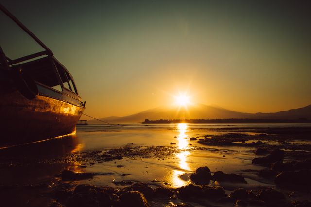 Old boat rests on shoreline with vibrant sunset in background creating warm golden reflections on water. Ideal for use in travel blogs, nature-themed presentations, relaxation and serenity illustrations, or promotional material for tourism industry showing tranquil beauty of natural landscapes.